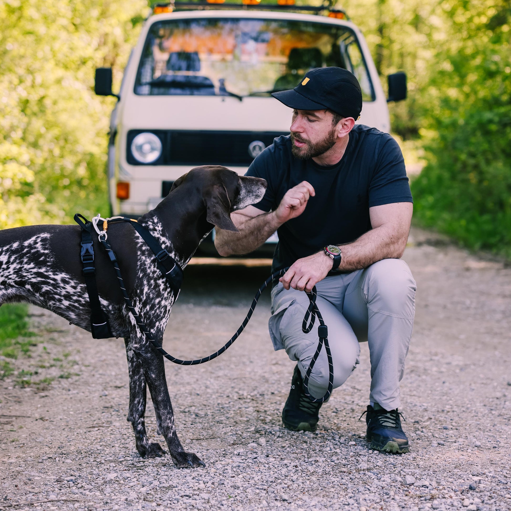 Man kneeling on a gravel road, petting a dog, with a beige van and trees in the background.