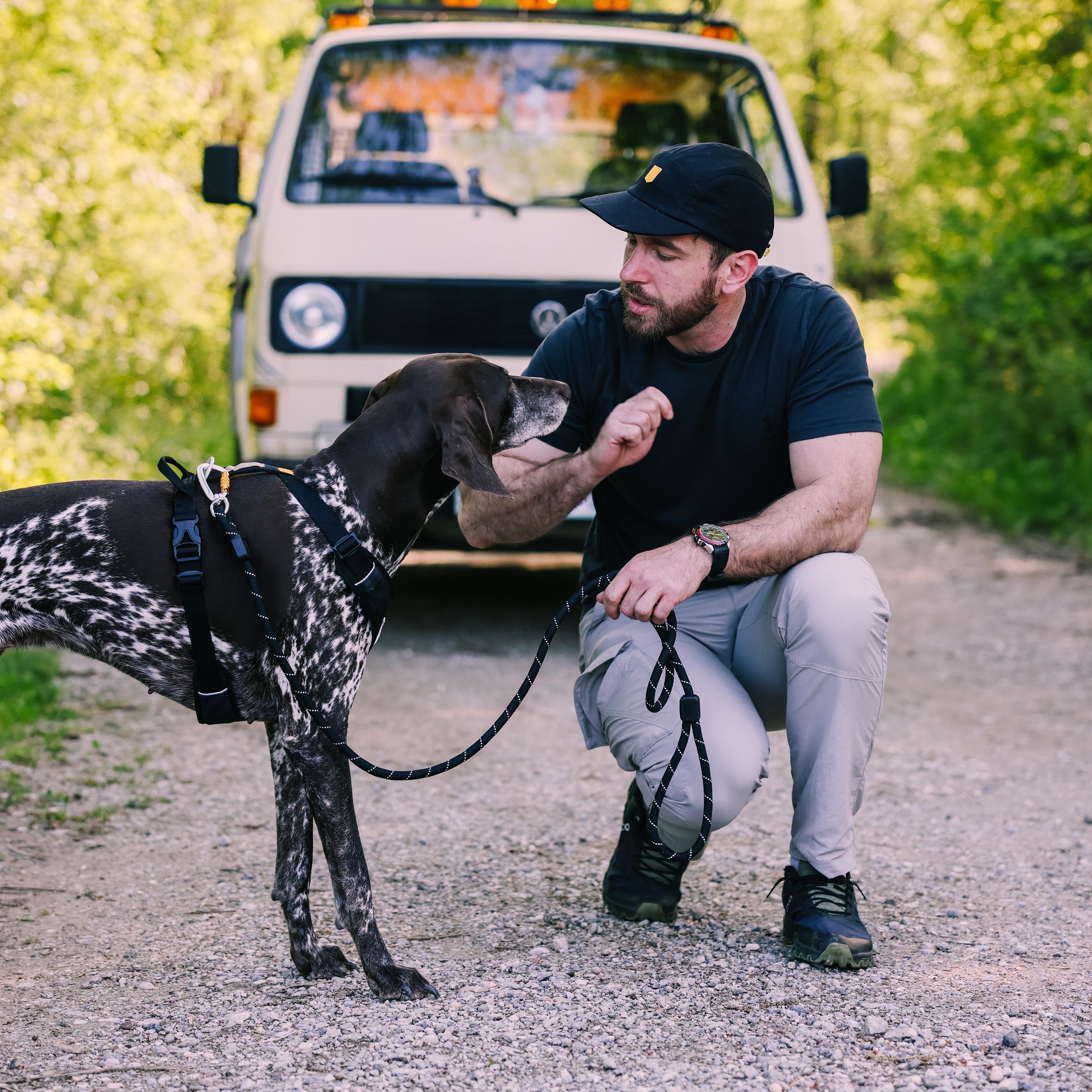 Man kneeling on a gravel road, petting a dog, with a beige van and trees in the background.