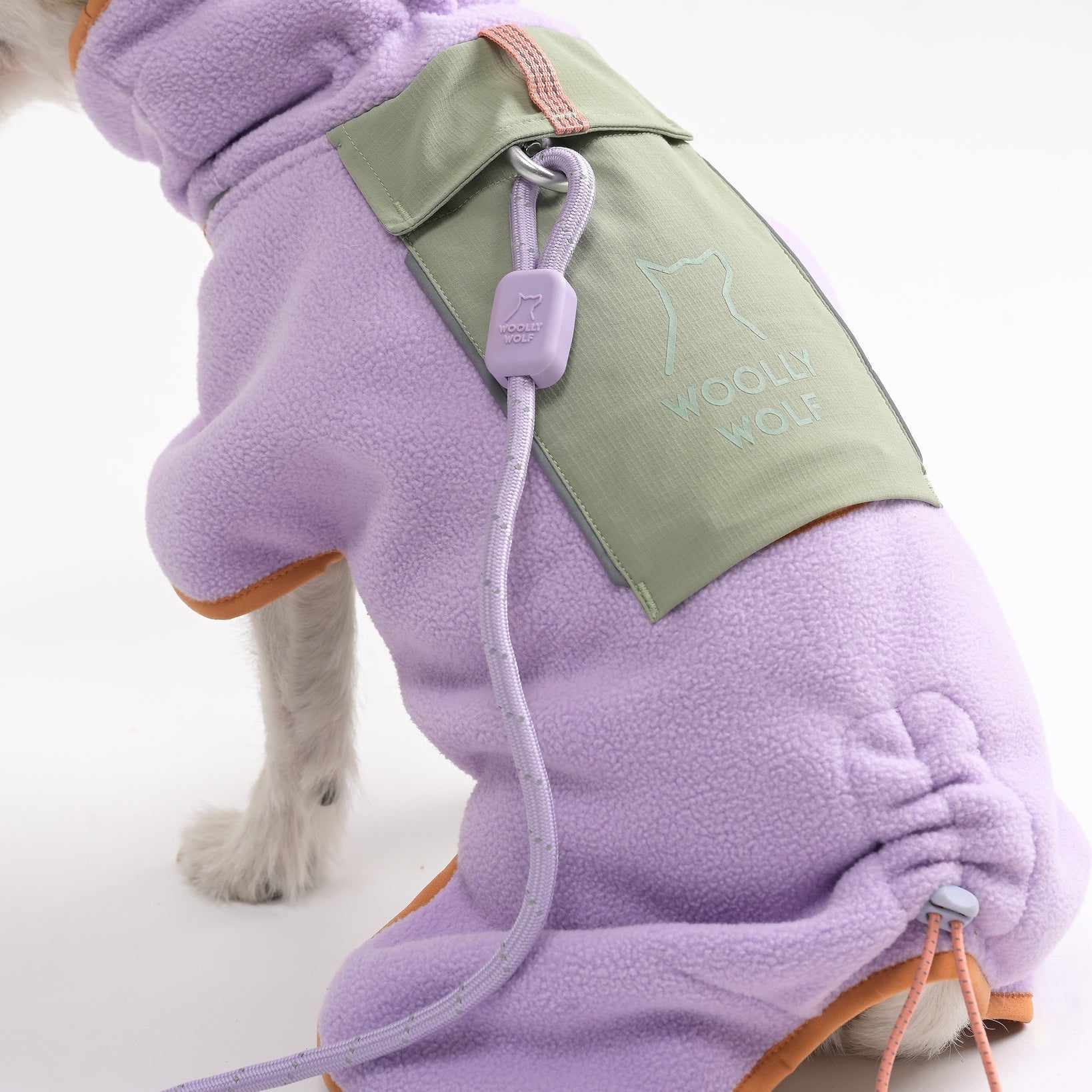 A small dog wearing a light purple fleece coat with orange trim and a green patch labeled Woolly Wolf. A matching purple leash attached to the coat is visible against a plain white background.
