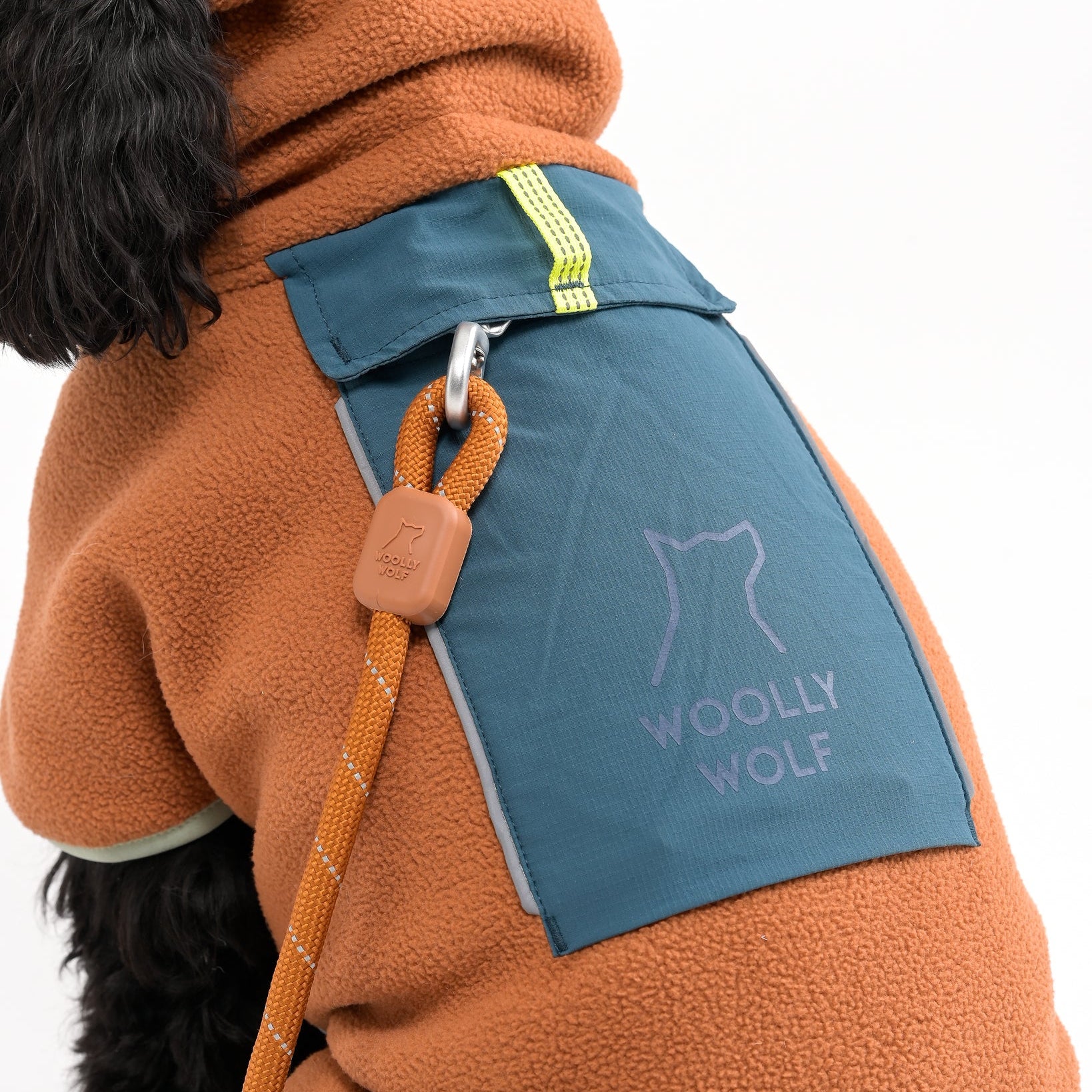 A dog wearing a brown fleece jacket with a blue pocket labeled WOOLLY WOLF and a leash attached to a metal ring on the jacket. The image is cropped to show only part of the dogs body.