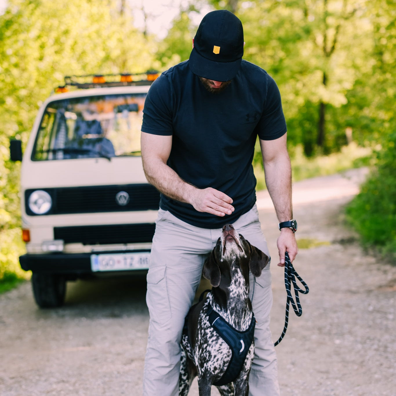 A man wearing an Alpha 360 Tech Cap Black and a t-shirt stands on a dirt path, giving a treat to a black and white harnessed dog. A white van is parked behind them among green trees.