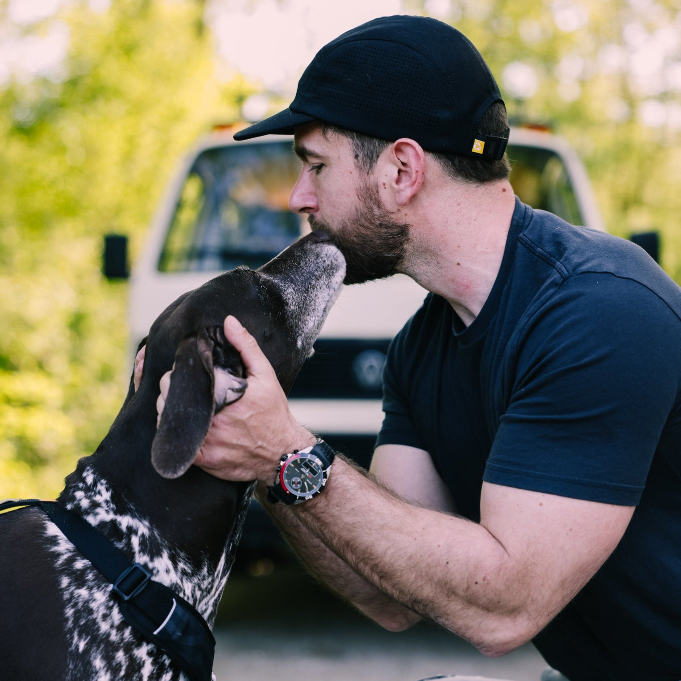 A man wearing the Alpha 360 Tech Cap Black kneels outdoors, gently holding and kissing the head of a black and white dog, with a white vehicle and greenery blurred in the background.