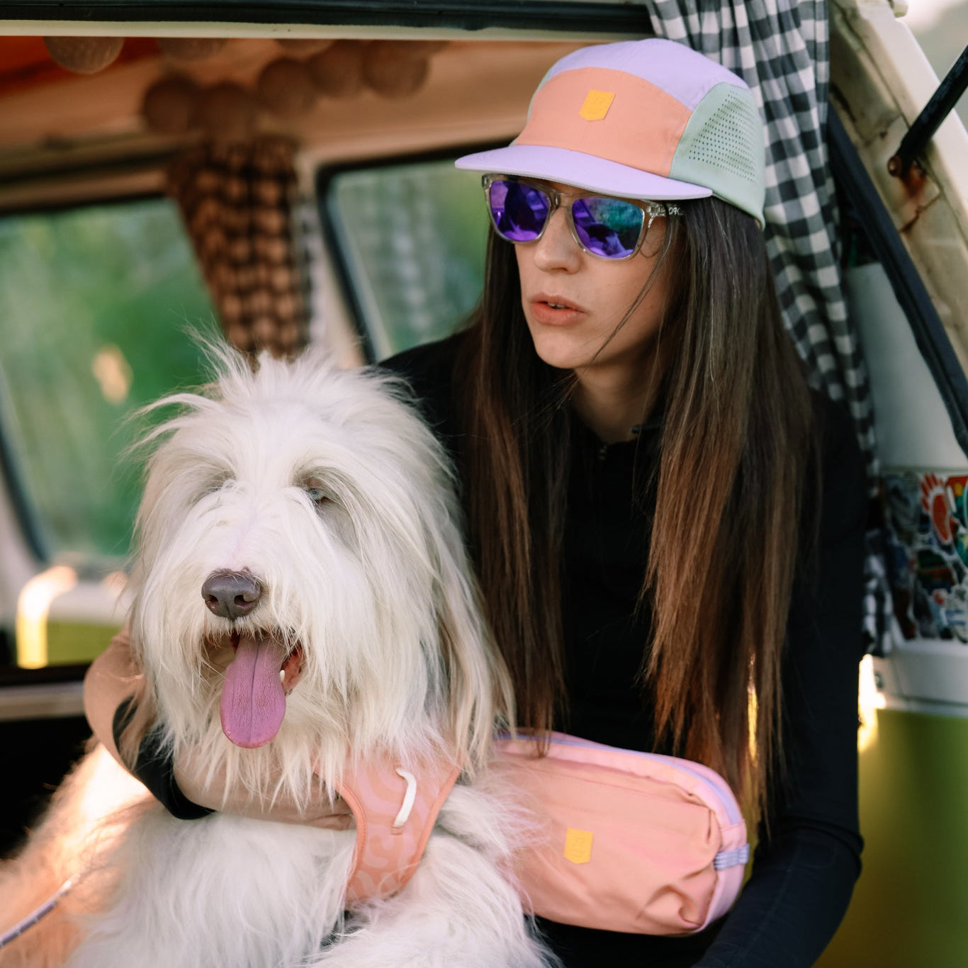 A woman in sunglasses wears the Alpha 360 Tech Cap Lavender and black clothing, sitting in a van with curtains and patterns. She holds a fluffy white dog with its tongue out and sports a pink fanny pack.