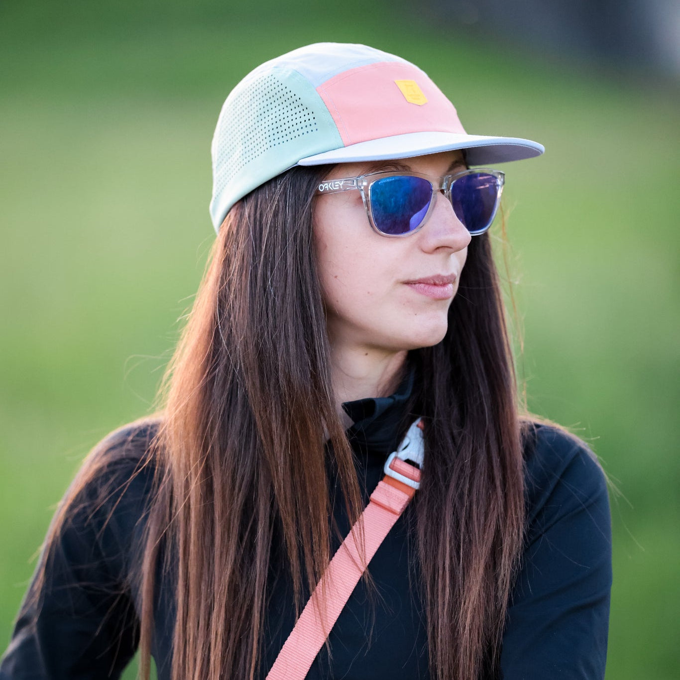 A woman with long brown hair wears the Alpha 360 Tech Cap in lavender, blue sunglasses, a black jacket, and a pink shoulder strap while standing outdoors against a blurred green grass background.