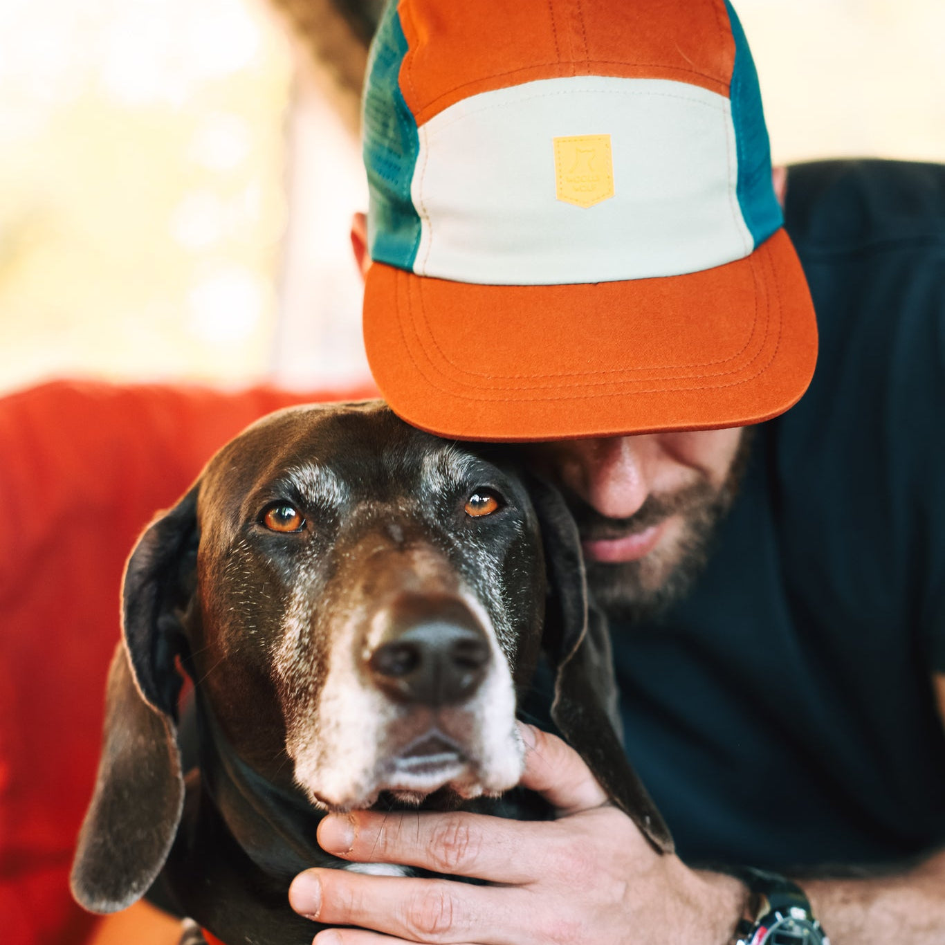 A man in an Alpha 360 Tech Cap Terracotta hugs a large black-and-white dog, gently holding its face. The cap partially hides his face, and both appear calm and close.