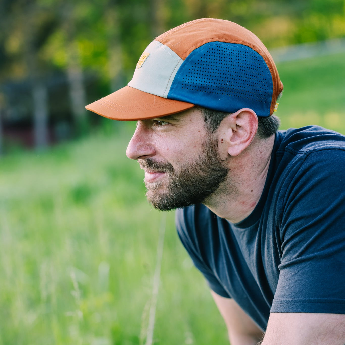 A bearded man wearing the Alpha 360 Tech Cap Terracotta and a dark t-shirt crouches in a grassy field, gazing thoughtfully to the left. Blurred trees and greenery fill the background.