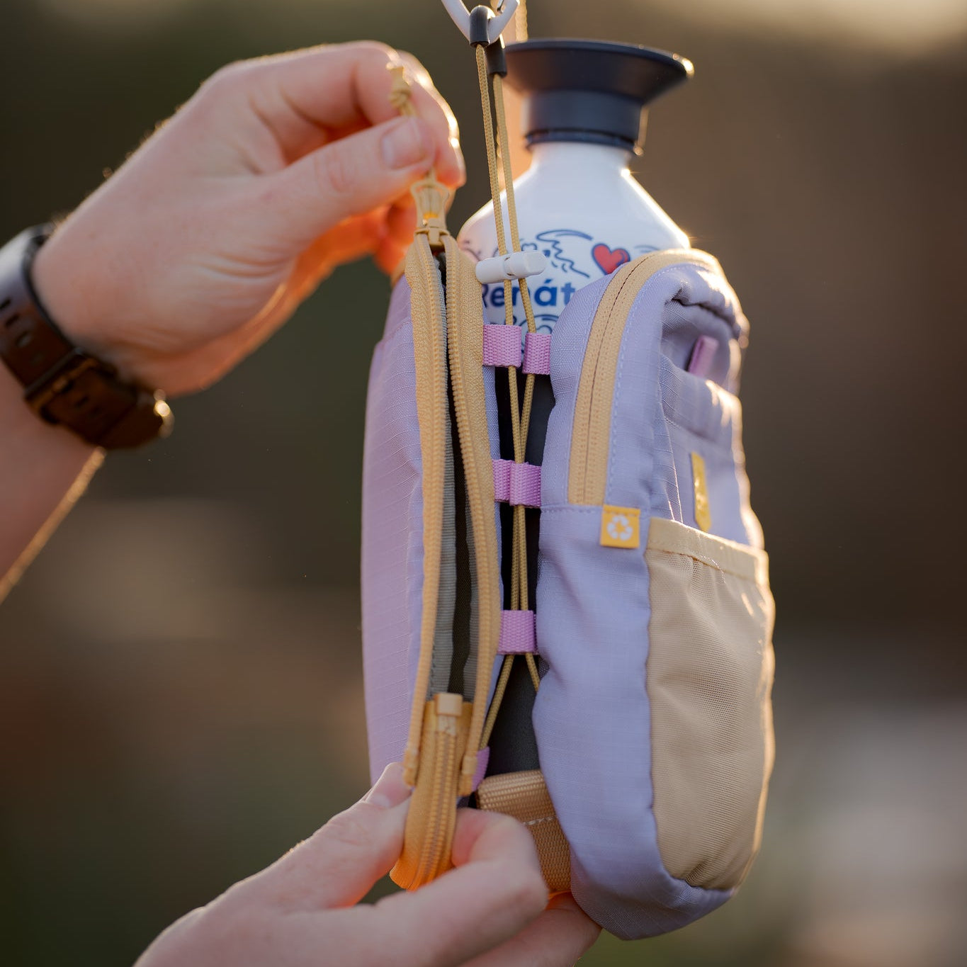 Person unzipping a small pastel bag with a water bottle inside, outdoors in soft sunlight.