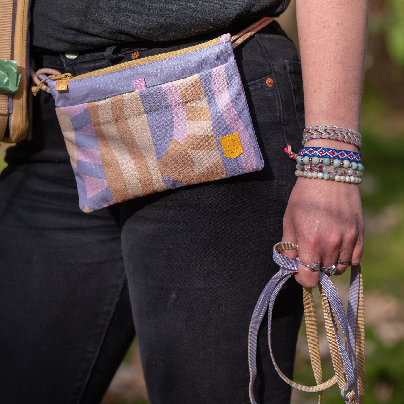 Person wearing black clothes with a pastel patterned fanny pack and holding woven leashes and beaded bracelets.