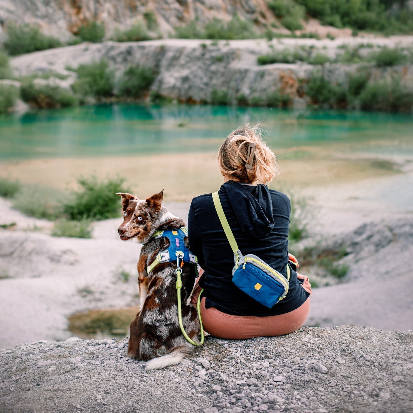 A woman with an Alpha 360 Hip Pack Deep Teal sits on a rock beside a dog, both facing a turquoise lake with rocky terrain and trees; the dog looks back at the camera.