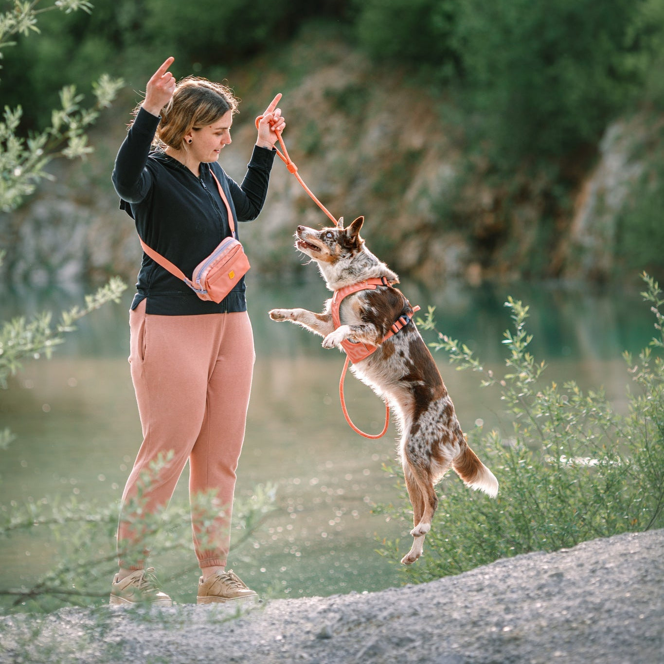 A woman wearing a black sweater and pink Alpha 360 Hip Pack holds a leash as her brown and white dog jumps beside her outdoors, with greenery and water in the background.