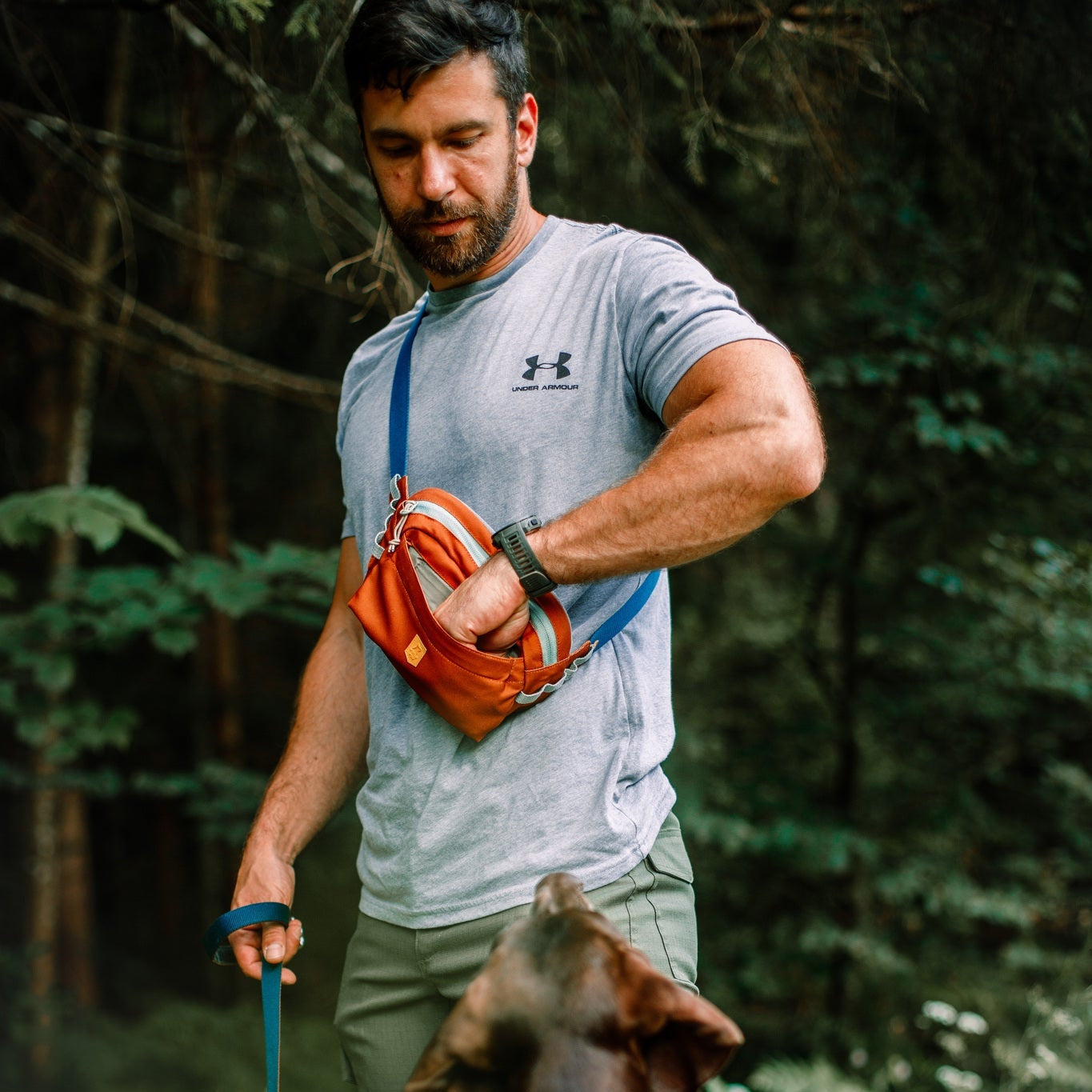 A man in a gray T-shirt opens the Alpha 360 Hip Pack Terracotta Mix while holding a leash, standing outdoors in a forest as his large dog looks up at him.