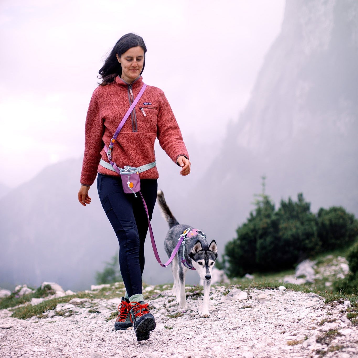 A woman in a red sweater walks a husky puppy on a rocky mountain trail, carrying the Alpha 360 Dog Snack Bag Mauve Mix, surrounded by misty mountains and lush greenery.