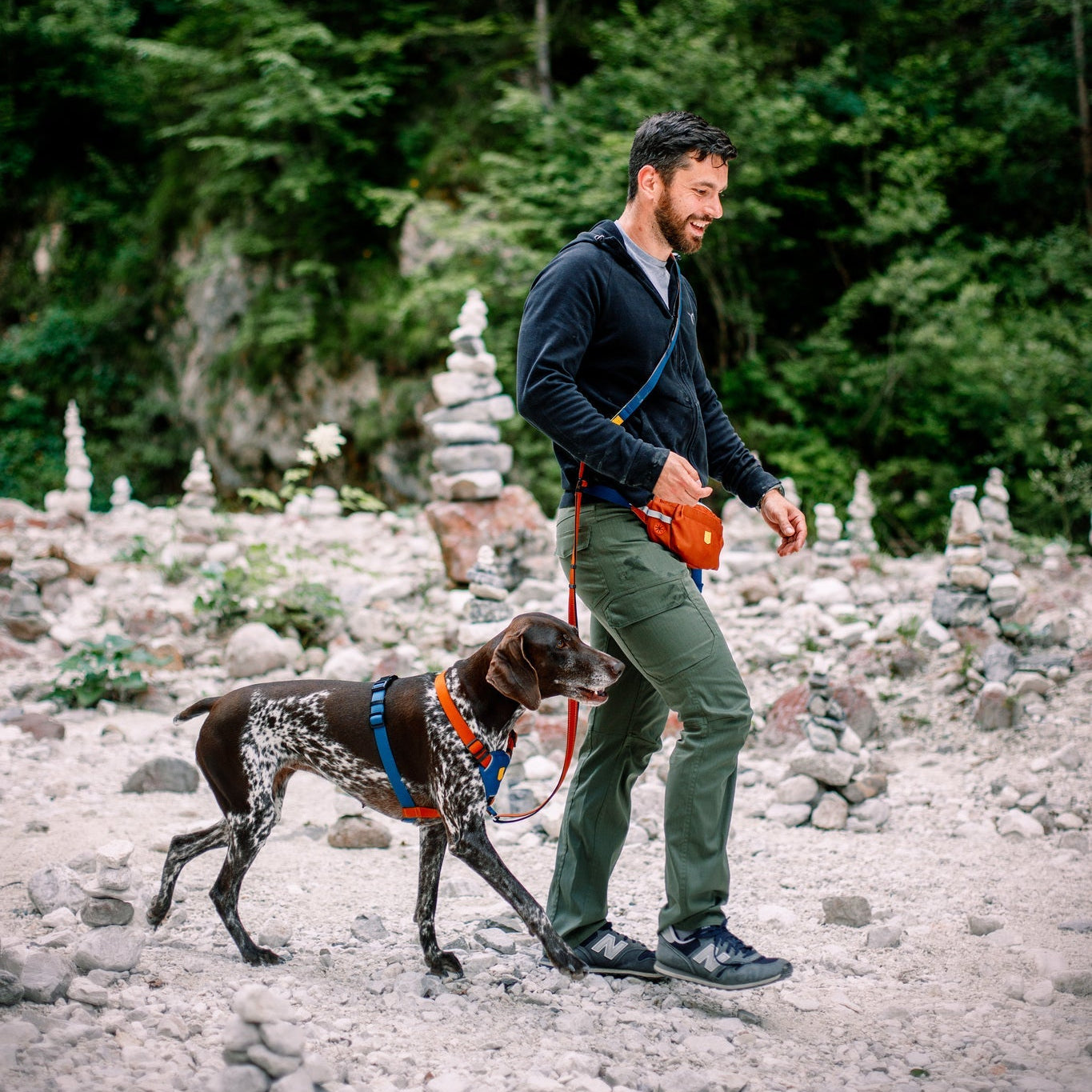 A man in a black sweater and green pants walks his brown and white dog, wearing a Color Block Dog Harness Lime Mix, on a leash along a rocky path with stacked stone piles and green trees in the background.