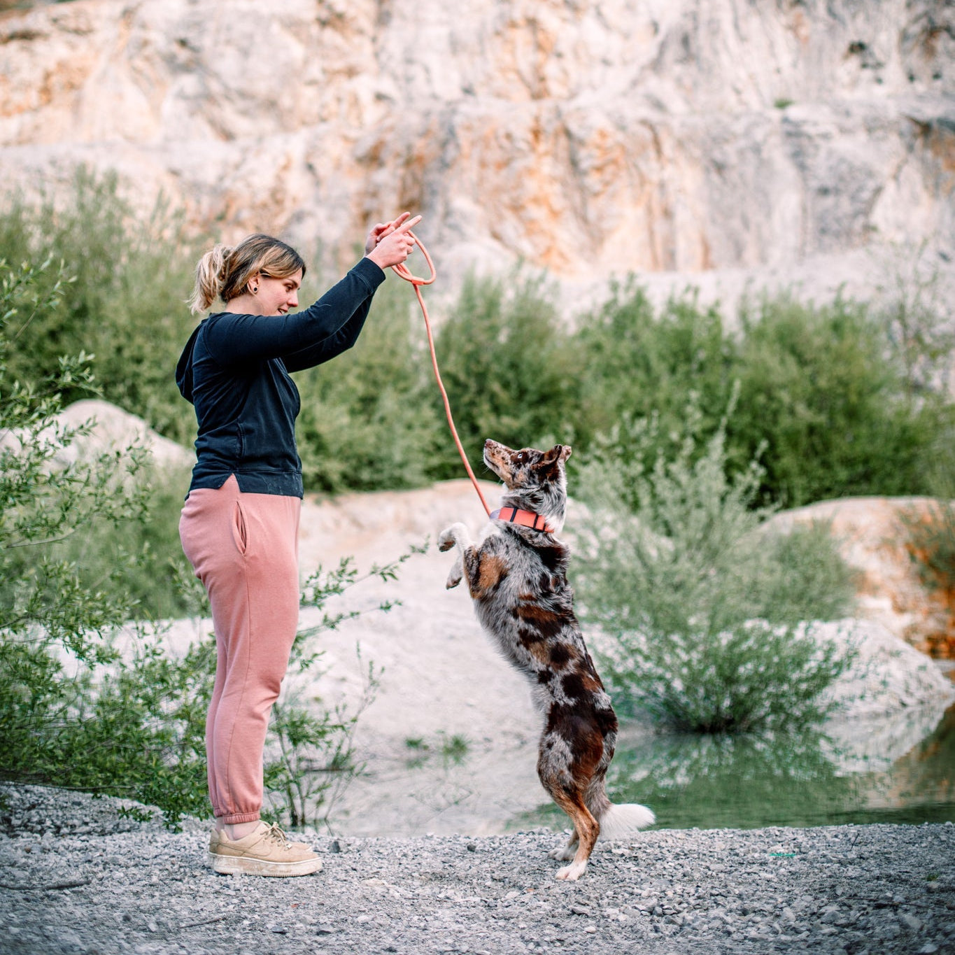 A woman in pink sweatpants holds up a leash attached to a Color Block Martingale Dog Collar Salmon Pink as her spotted dog stands on its hind legs, with rocky hills and green bushes in the background.
